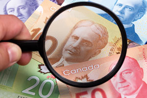 Various Canadian currency bills are spread out and a person holding a magnifying glass is inspecting one of them.