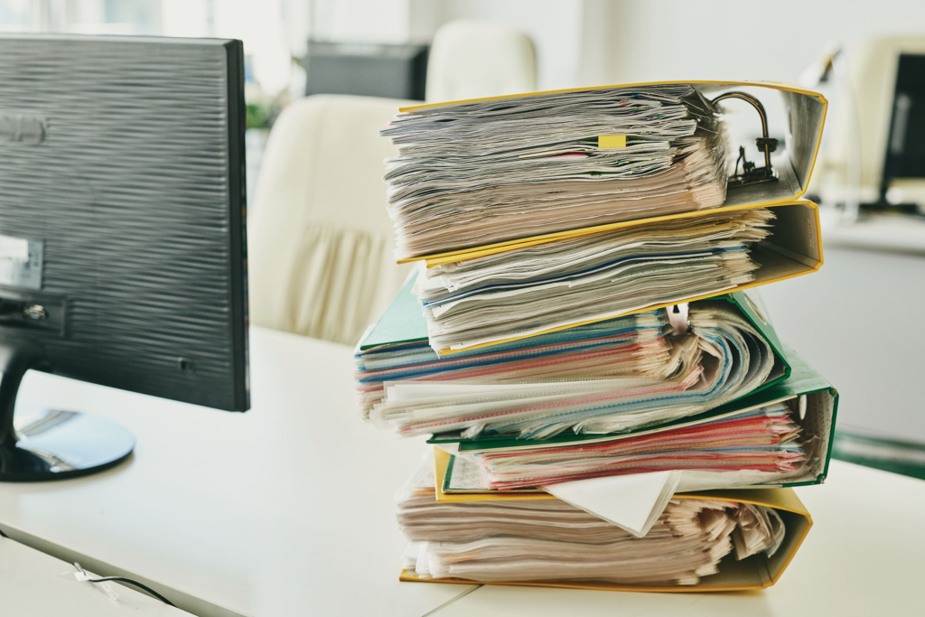 Thick binders full of paper are stacked on top of each other on a desk.