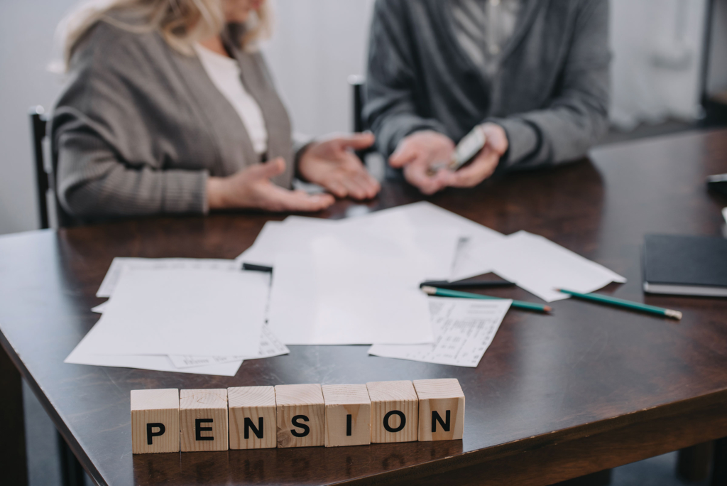 Two people sit at a table with paper in front of them. Wooden blocks on the table spell the word pension.